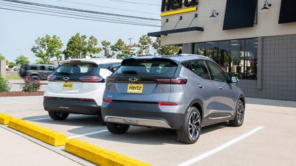 A Summit White 2023 Chevrolet Bolt EV and a Gray Ghost Metallic 2023 Chevrolet Bolt EUV parked in front of a Hertz rental location in Michigan. 