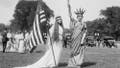 A woman holds up an American Flag while wearing a white dress. Her companion wears a Statue of Liberty costume. Photo circa 1919.