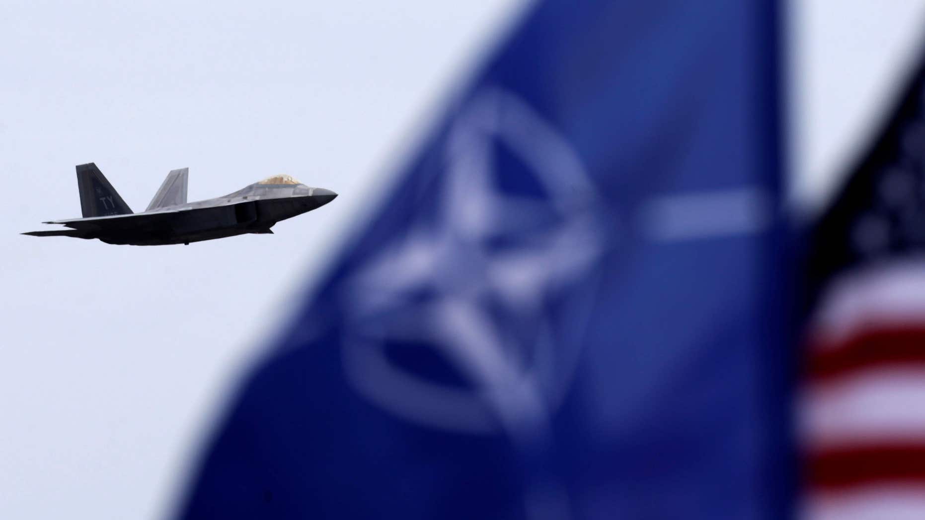 April 27, 2016: NATO and U.S. flags flutter as U.S. Air Force F-22 Raptor fighter flies over the military air base in Siauliai, Lithuania.