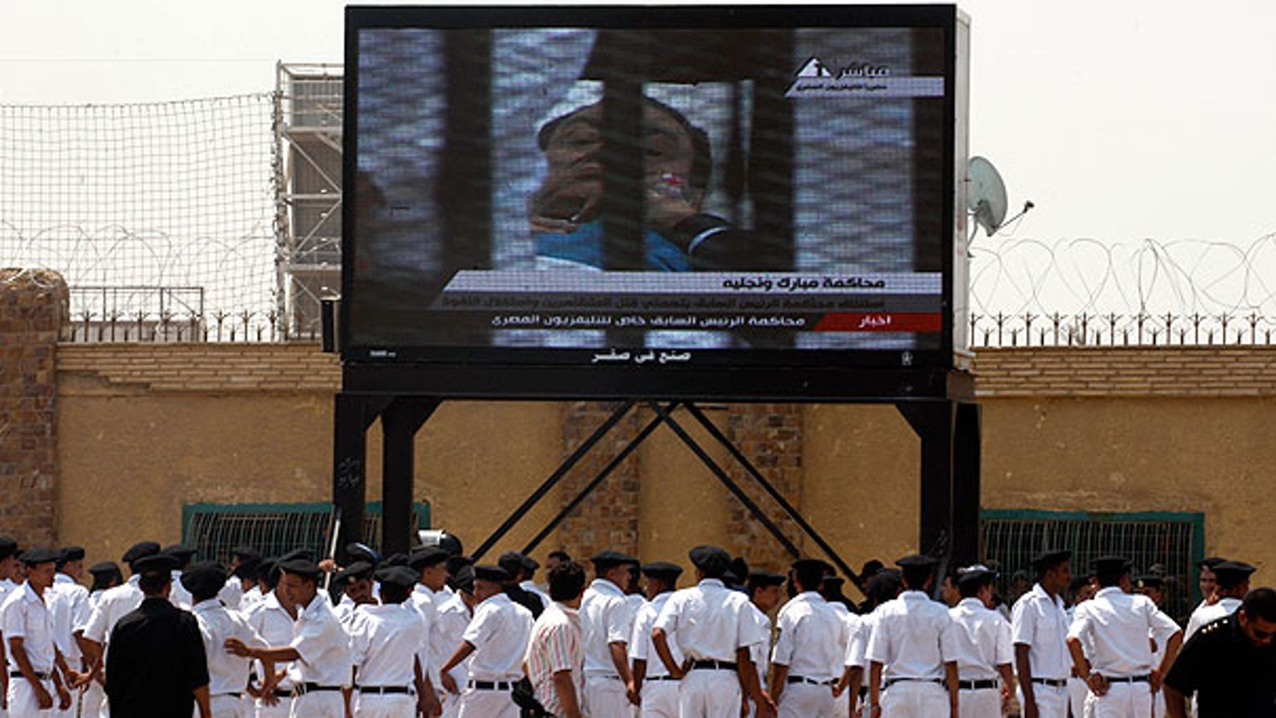 Aug. 15: Egyptian police officers watch 83-year-old former Egyptian president Hosni Mubarak on a bed in the defendants' cage from the courtroom live on a large TV screen outside the Police Military Academy complex in Cairo, Egypt.