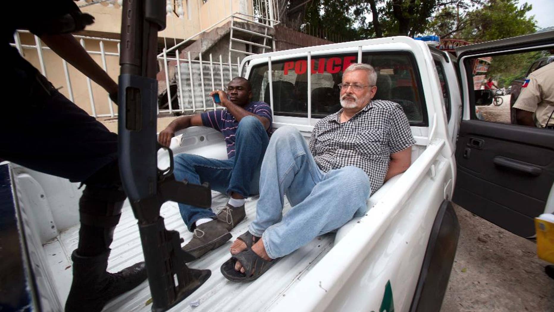 FILE - In this Friday, Sept. 5, 2014, file photo, Michael Geilenfeld waits in handcuffs as the manager of his orphanage sits with him in the back of a police truck outside the St. Joseph's Home For Boys after police closed it down in the Delmas area of Port-au-Prince, Haiti. Hearts With Haiti, a nonprofit that raises money for the orphanage, and Geilenfeld have accused activist Paul Kendrick of spreading falsehoods that have cost the charity more than $2 million in donations. A jury that was seated Monday, July 6, 2015 for the trail. (AP Photo/Dieu Nalio Chery, File)