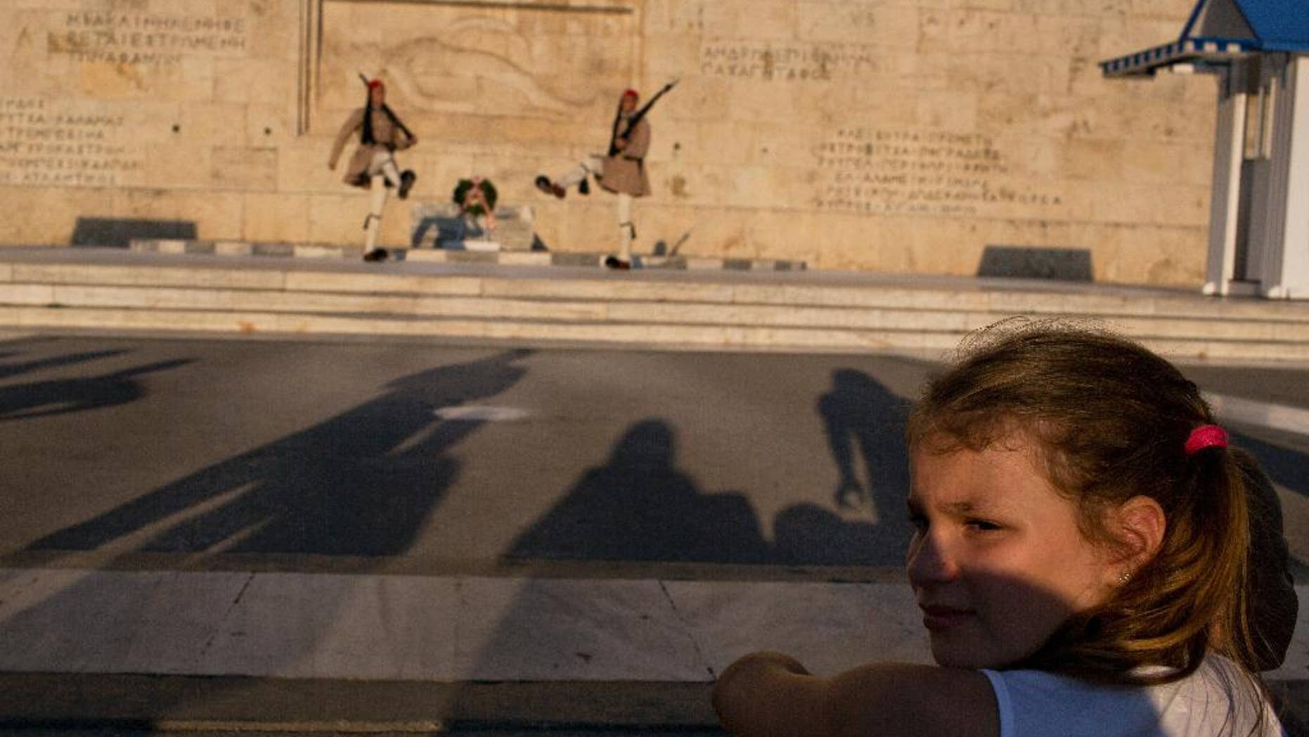 A visitor looks away from the changing of the guard ceremony at Greece's parliament to watch a small number of protesters across the street, in central Athens on Tuesday, July 5, 2016. The tiny anti-government rally was held to mark a year since Greeks voted against bailout measures in a referendum. Shortly after the vote, Greece reaching a third bailout deal with international rescue creditors. (AP Photo/Petros Giannakouris)