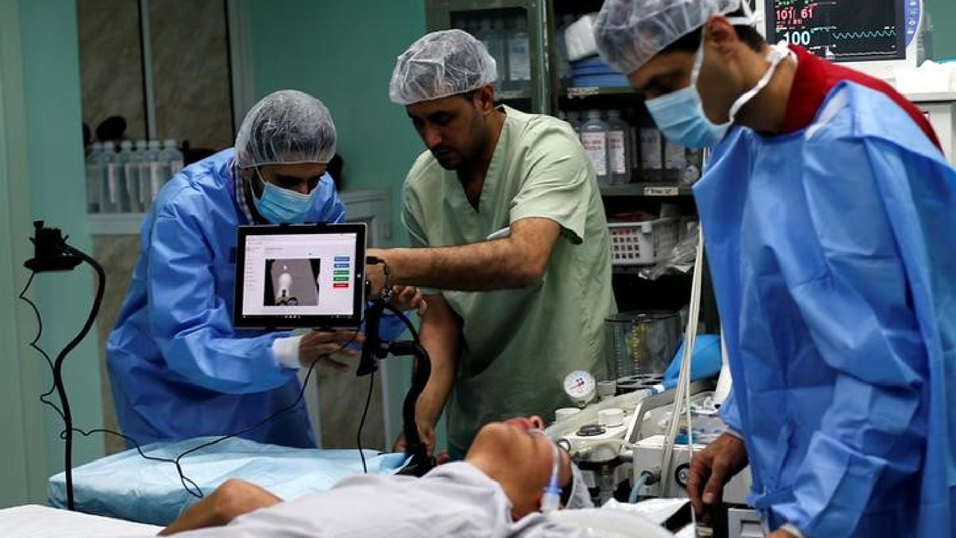 A patient lies on a bed as a technician adjusts a screen before the start of a Proximie surgery, with doctor Ghassan Abu Sitta in Beirut guiding Palestinian surgeon Hafez Abu Khousa in the operating room at Al Awda Hospital in the northern Gaza Strip
