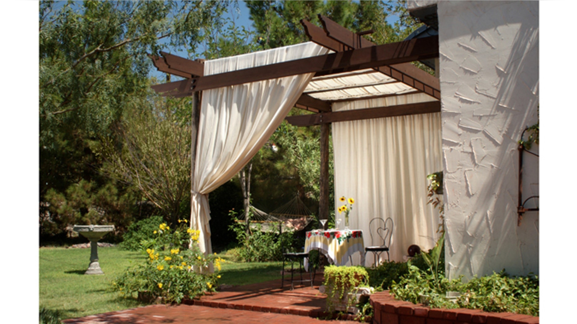A brick patio with wooden patio posts and a cover of canvas curtains. The back ground has green grass, a birdbath and hammock.