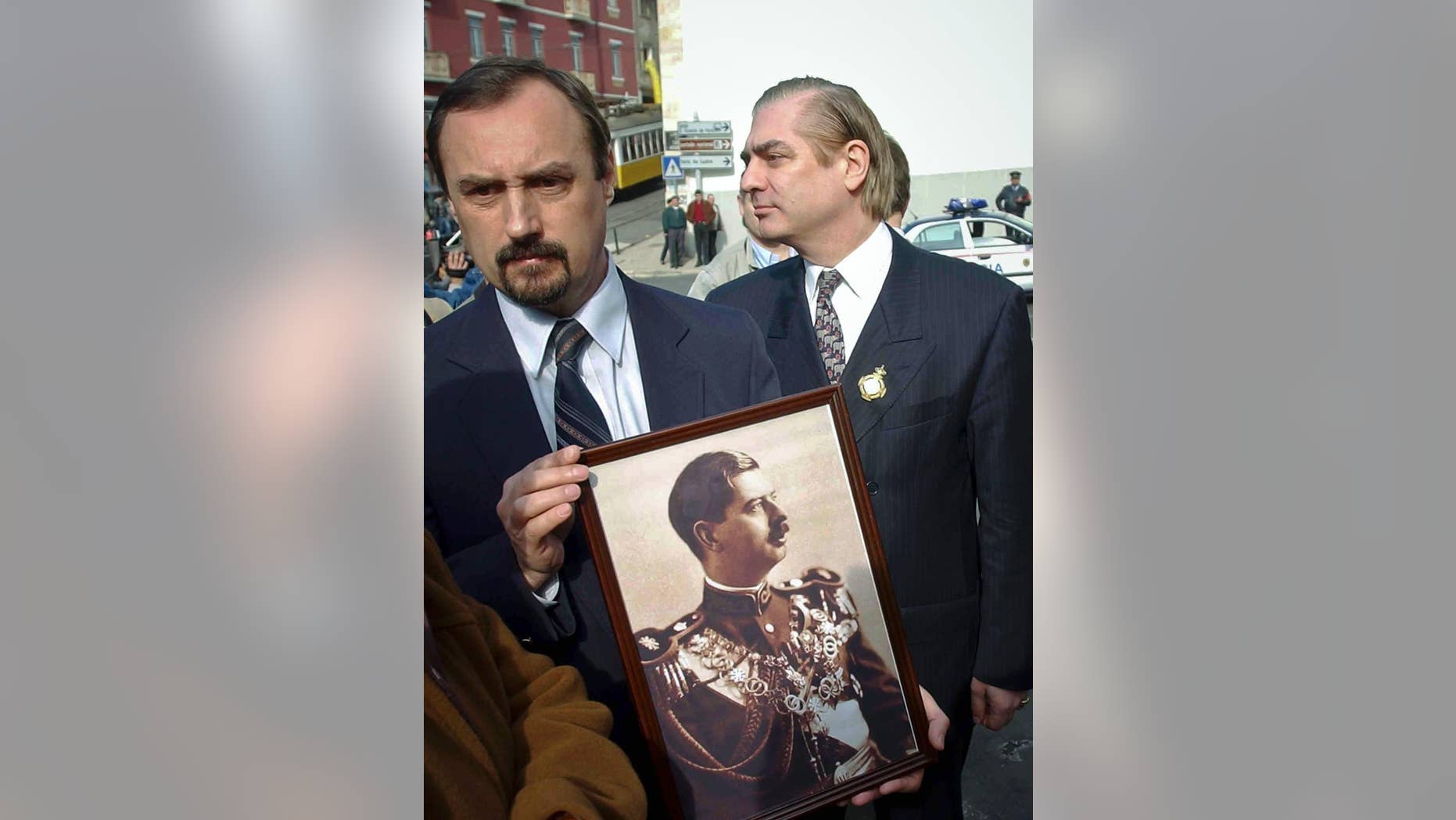 FILE - In this Feb.13 2003, file picture an unidentified man holds a photograph of Romania's King Carol II as Romania's Prince Paul, right, walks past outside a church in Lisbon, Portugal, where the king was buried. Prosecutors have detained Romania's Prince Paul, Friday, Dec. 11, 2015, in a case of alleged real estate fraud involving a top aide to a former prime minister and a newspaper editor.(AP Photo/Armando Franca, File)