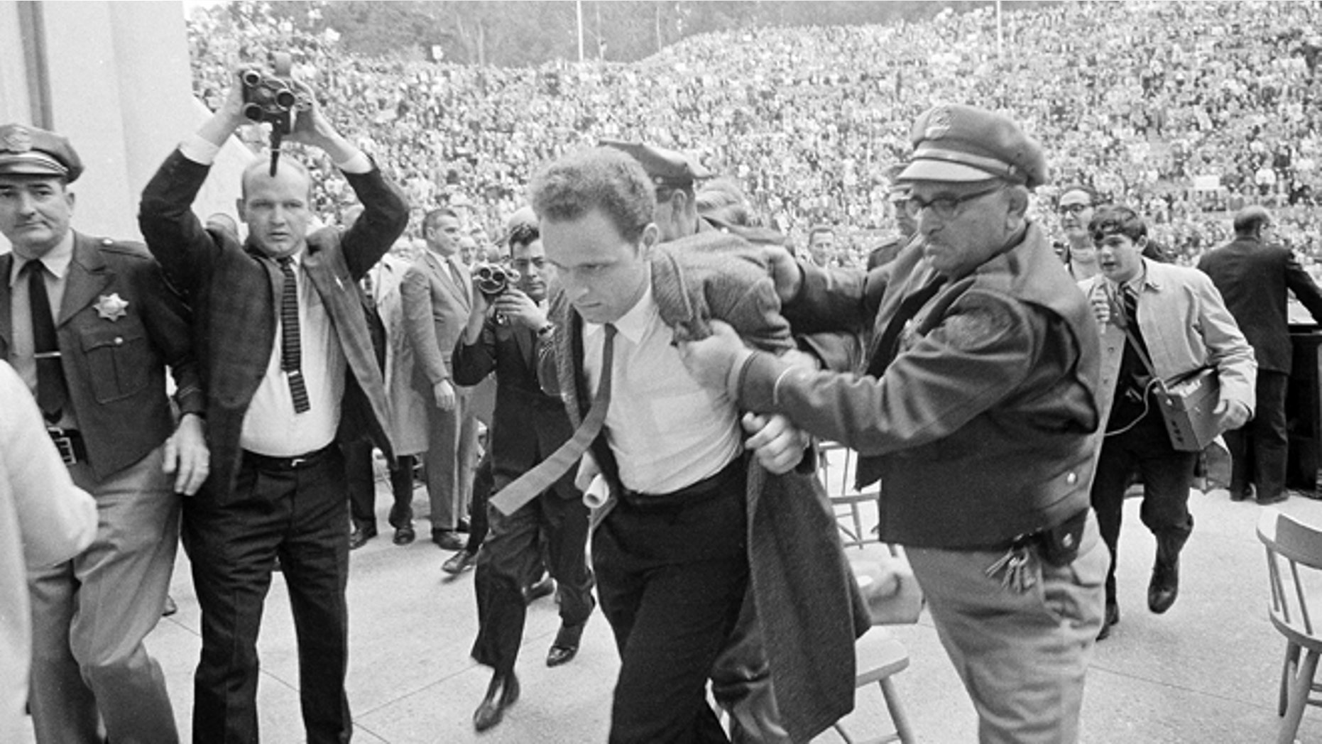 FILE 1964: Mario Savio, leader of the Berkeley Free Speech Movement, is restrained by police as he walks on to the platform at the University of California's Greek Theater in Berkeley, Calif. Savio attempted to speak directly following the appearance of University President Clark Kerr. He was later permitted to make two announcements to the assembled students.