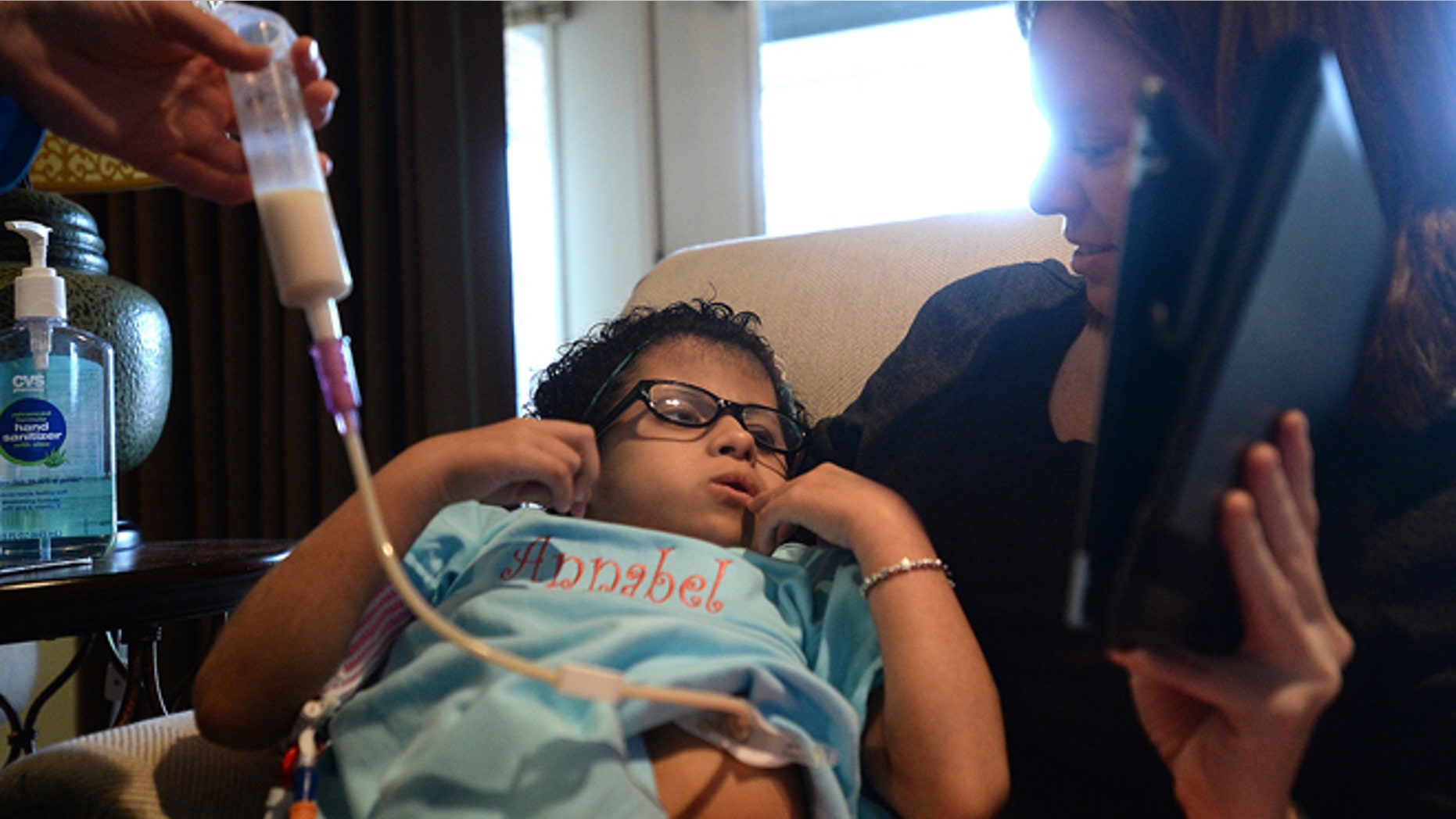 Annabel Shelander watches an episode of Barney with older sister Tara as nurse aide Megan Goats gives her a feeding through her peg tube at the family's home in Beaumont, Texas, on Saturday, Feb. 14, 2015. Shelander, 9, has a rare condition that keeps her from consuming and digesting food naturally. (AP Photo/The Beaumont Enterprise, Kim Brent)