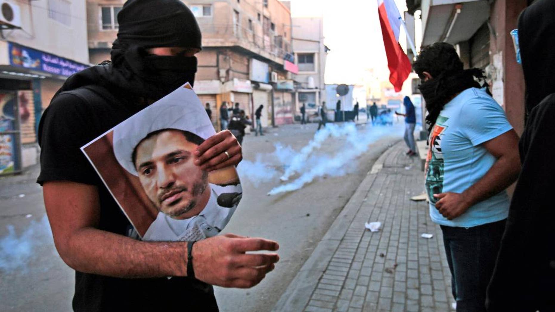 FILE- In this Jan. 3, 2015 file photo, a masked Bahraini anti-government protester holds a picture of jailed Shiite cleric Sheik Ali Salman, the head of the opposition al-Wefaq political association, as riot police fire tear gas canisters during clashes in Bilad Al Qadeem, Bahrain. On Tuesday, June, 14, 2016, Bahrain said it has suspended all activities by Al-Wefaq, the largest Shiite opposition political group, and frozen its assets amid a widening crackdown on dissent, five years after the country’s Arab Spring protests. A statement from the Justice and Islamic Affairs Ministry carried on the state-run Bahrain News Agency said a court made the decision to suspend Al-Wefaq to “safeguard the security of the kingdom.” (AP Photo/Hasan Jamali, File)