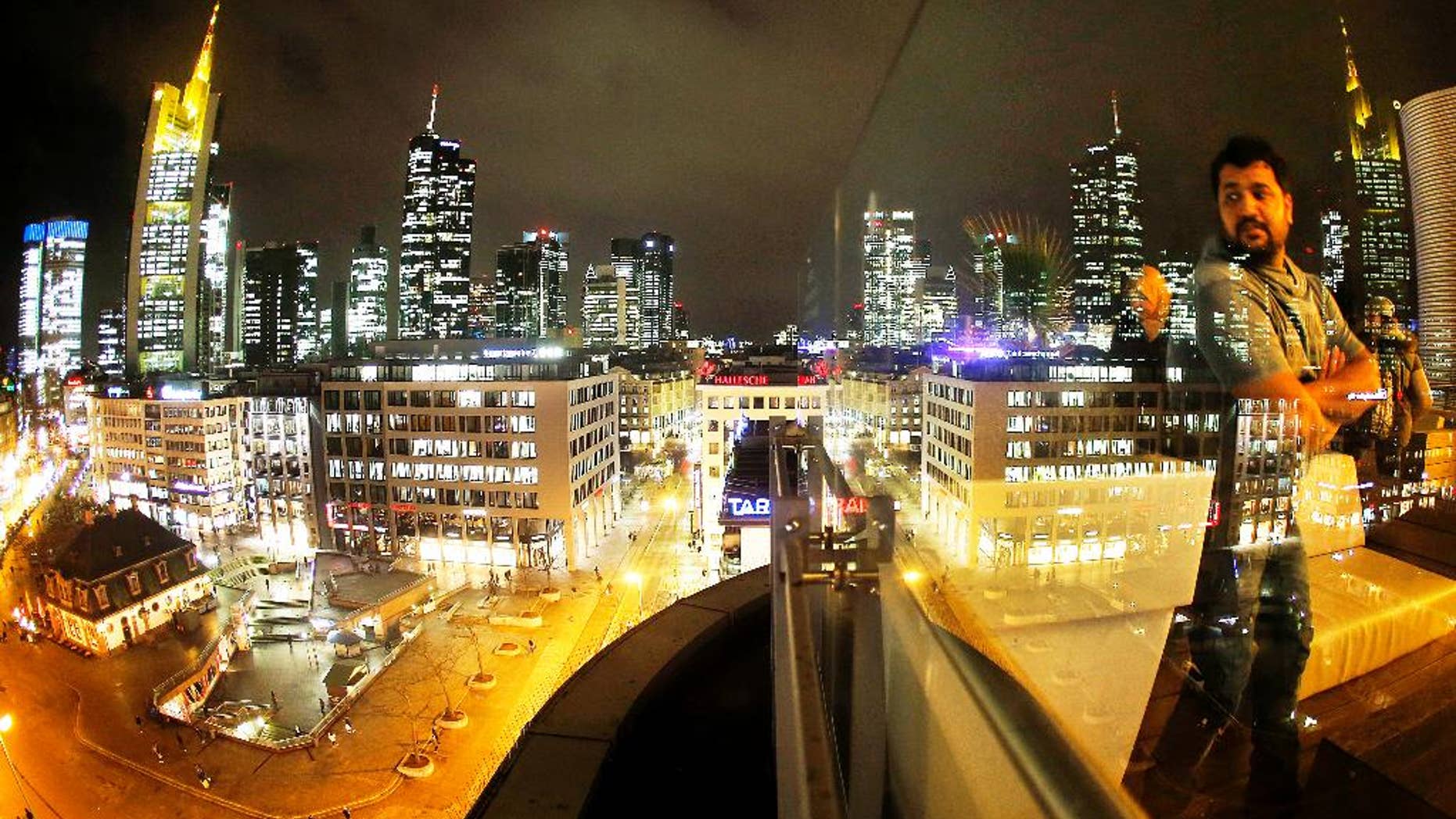The buildings of the banking district are reflected in a glass railing while a man watches from a skyline outdoor restaurant in Frankfurt, Germany, Wednesday, Jan. 4, 2017. (AP Photo/Michael Probst)