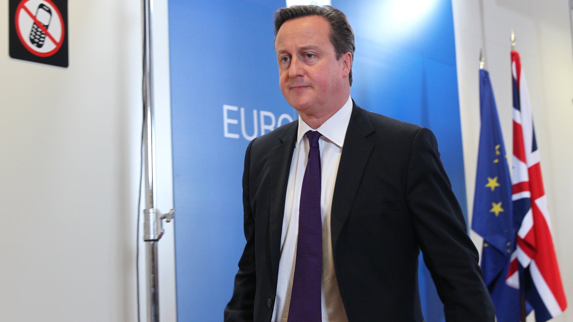 British Prime Minister David Cameron walks past a sign prohibiting the use of mobile phones, after he addressed the media at the European Council building in Brussels, Friday, Oct. 25, 2013. Migration, as well as an upcoming Eastern Partnership summit, tops the agenda in Friday's meeting of EU leaders. (AP Photo/Yves Logghe)
