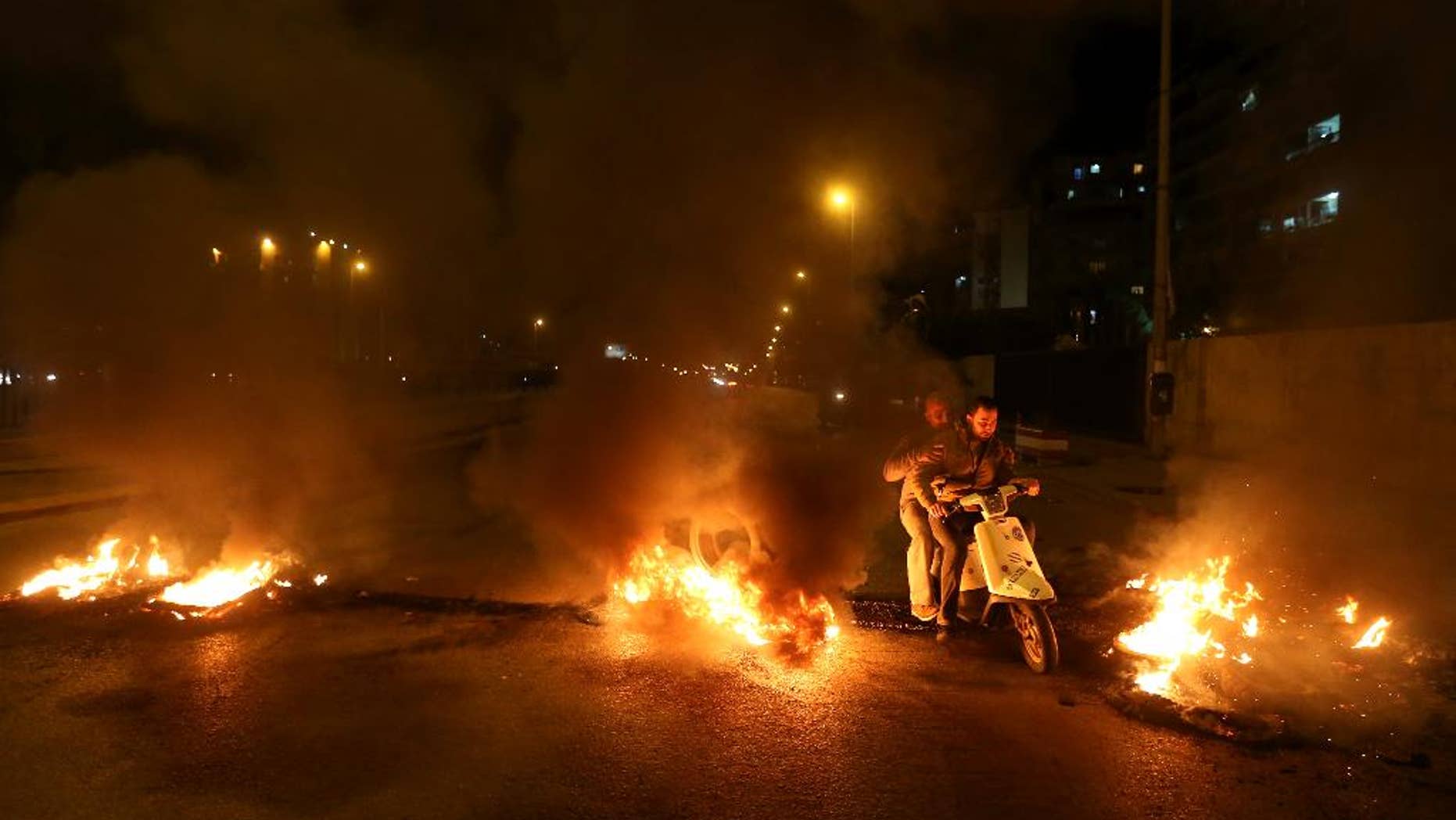 Lebanese citizens ride their scooter between burning tires that were set on fire by Sunni protesters, at one of the major roadways between Beirut and the capital's southern suburbs, a Hezbollah stronghold, to show their support with Sunni town of Arsal, in Beirut, Lebanon, Tuesday, March 18, 2014. Gunmen from Lebanon's militant Hezbollah group and local Shiite Muslim residents tightened their blockade of a Sunni town of Arsal near the Syrian border Tuesday, sparking concerns that thousands of Syrian refugees stranded in the area could be cut off from humanitarian aid. (AP Photo/Hussein Malla)