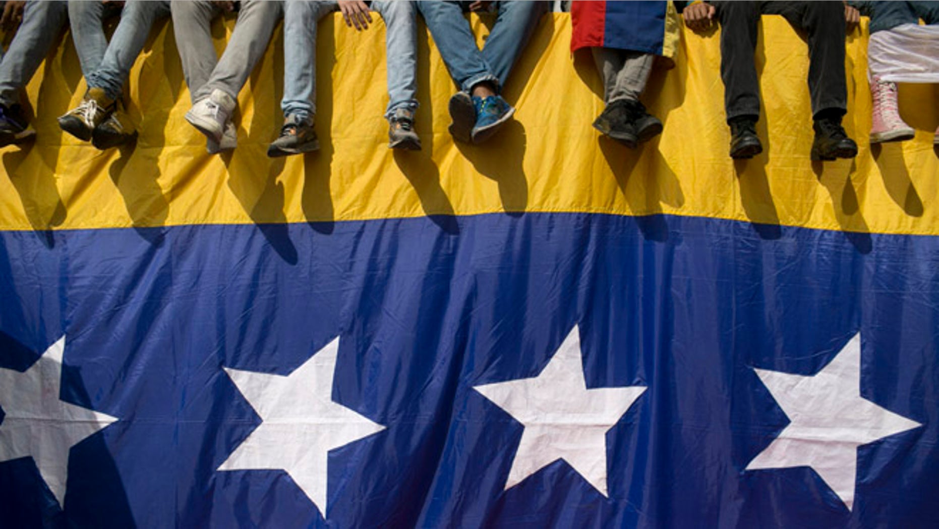 People sit on a wall draped by a Venezuelan flag during a protest against President Nicolas Maduro in Caracas, Venezuela, Wednesday, Oct. 26, 2016. Venezuela's standoff deepened after congress voted to open a political trial against Maduro for breaking the constitutional order and opposition leaders called for mass demonstrations on Wednesday to drive the leader from office. (AP Photo/Alejandro Cegarra)