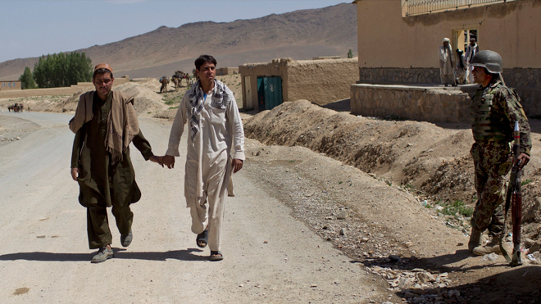 May 17, 2012, Two men walk hand-in-hand past an Afghan National Army soldier in east Afghanistan.