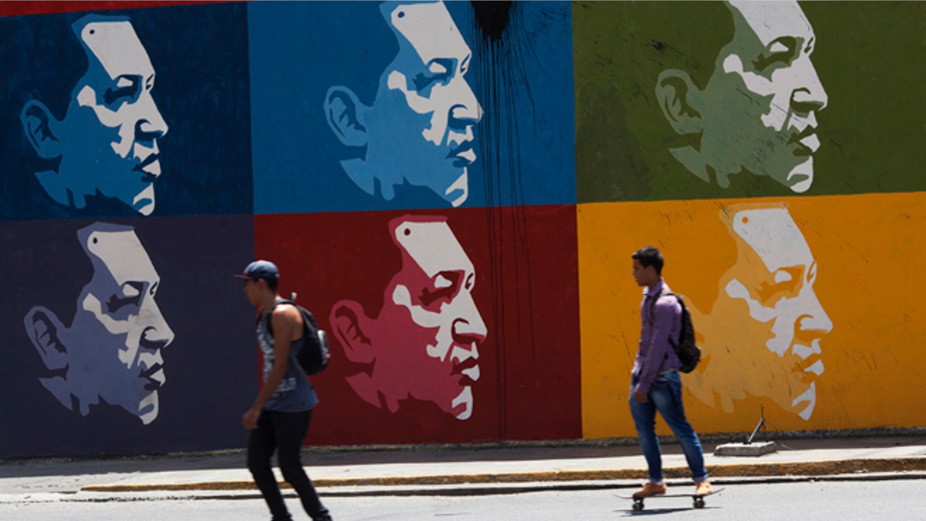 Two men skate board past a mural of Venezuela's late President Hugo Chavez in downtown Caracas, Venezuela, Tuesday, March 4, 2014. Hes been dead a year, but Chavezs face and voice are everywhere. On Wednesday, the country will mark Chavez's death, who died at age 58 on March 5, 2013. (AP Photo/Rodrigo Abd)