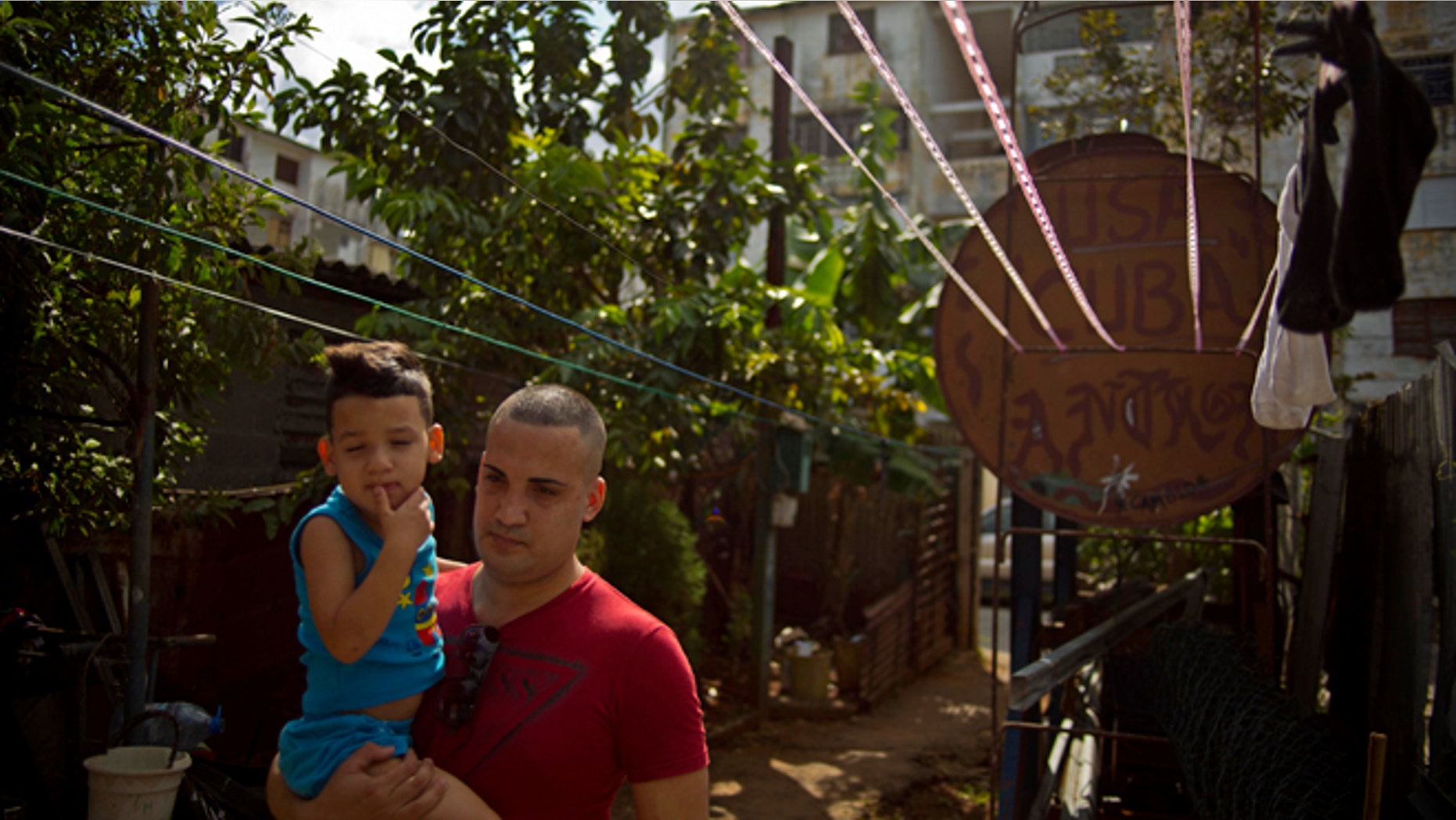 A day after his release, dissident Miguel Alberto Ulloa, 25, holds his son, as he walks beside a water tank painted with the acronym for the United States and his country's name, outside his home, in Havana, Cuba, Friday, Jan. 9, 2015. Advocates, relatives and dissidents say that Cuba has freed at least 38 people on a U.S. list of imprisoned opposition members. U.S. officials said Friday after weeks of virtual silence that those released had been on the list of 53. (AP Photo/Ramon Espinosa)