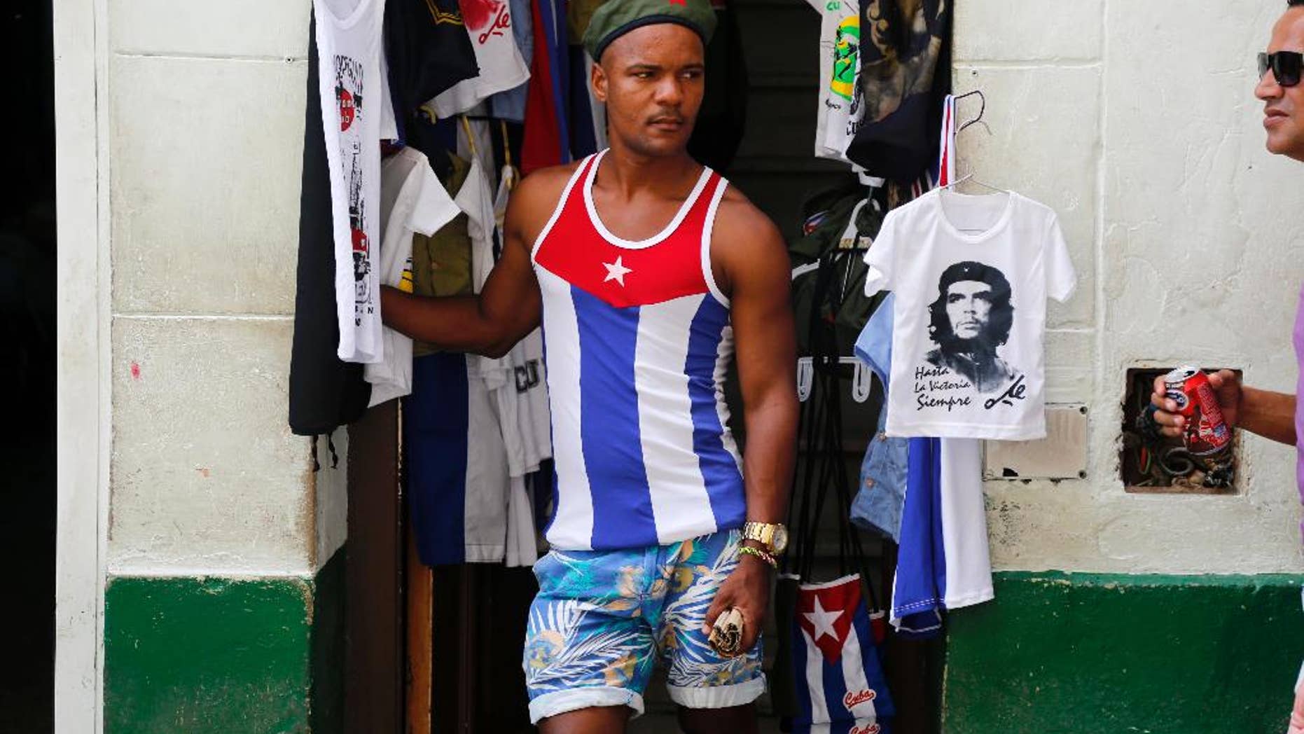 In this June 1, 2015 photo, a vendor, wearing a muscle shirt with a Cuban national flag motif, stands next to a souvenir T-shirt featuring Che Guevara, in the shop's doorway, in Old Havana, Cuba. Visitors who wander through Old Havana or Habana Vieja, as locals call it, will find that every street seems to have a sign attesting to something of cultural or historic significance. (AP Photo/Desmond Boylan)