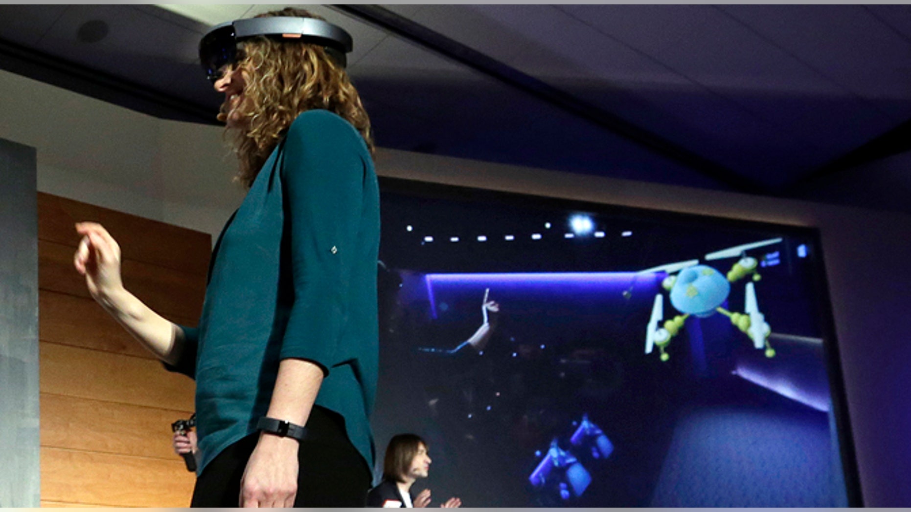 In this photo taken Wednesday, Jan. 21, 2015, Microsoft's Lorraine Bardeen demonstrates a hologram device as what she "sees" is projected on a screen behind during an event at the company's headquarters in Redmond, Wash. With the new HoloLens headset, Microsoft is offering real-world examples to show how you might use three-dimensional digital images _ or holograms _ in daily life. (AP Photo/Elaine Thompson)