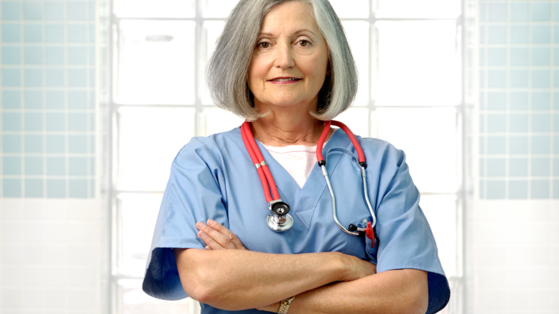 senior female doctor stands with her arms crossed and wears blue scrubs with a red stethoscope around her neck