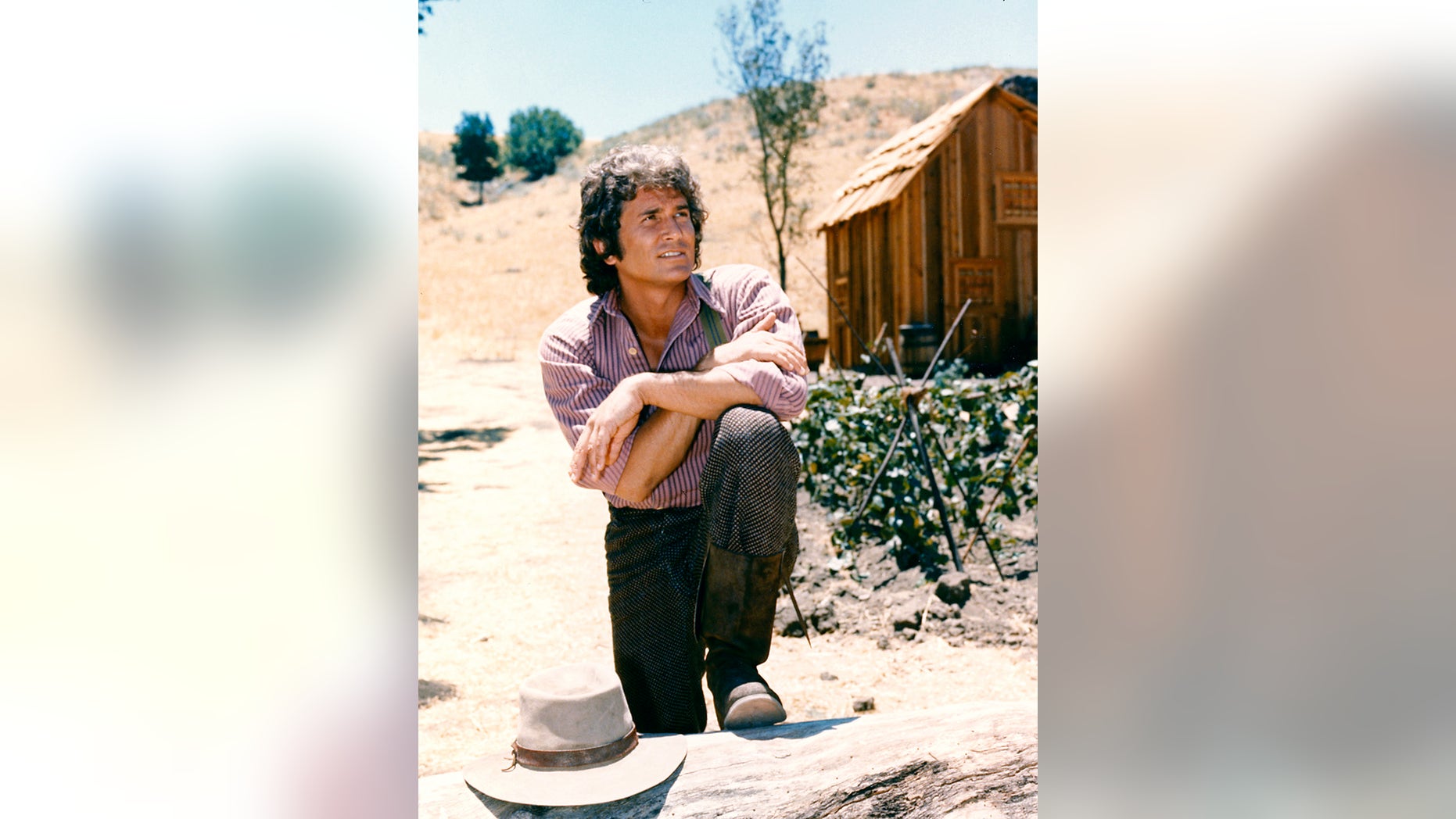 Michael Landon (1936-1991), US actor, poses with his arms crossed and resting on his knee with a log cabin the background in a portrait issued as publicity for the US television series, 'Little House on the Prairie', circa 1974. The drama, adapted from the novels by Laura Ingalls Wilder (1867-1957), starred Landon as 'Charles Ingalls'.