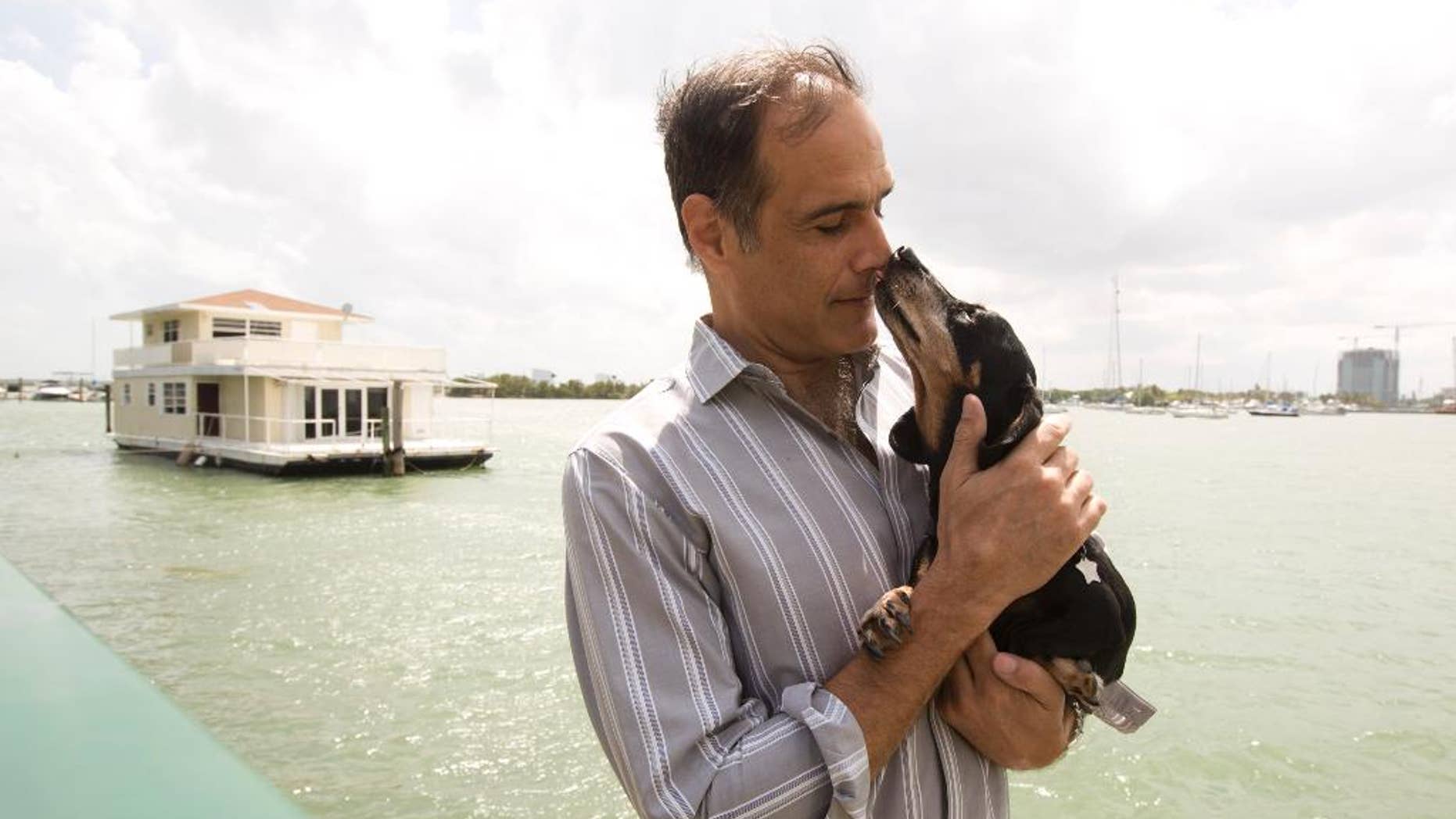 In this April 22, 2014 photo, Fane Lozman poses for photos holding his dog and in front of his home floating in the waters near North Bay Village, Fla. He caught legal lightning in a bottle last year when the U.S. Supreme Court agreed with him that his floating home was a house, not a vessel covered by maritime law. But the justices haven’t had the last word: Lozman is still fighting for compensation for the home, which was destroyed years ago. The Fort Lauderdale-based federal judge whose decision on the floating home was overturned, U.S. District Judge William Dimitrouleas, refused earlier this year to give Lozman any of the $25,000 bond posted by the city of Riviera Beach to pay for Lozman’s home in case he won. (AP Photo/J Pat Carter)