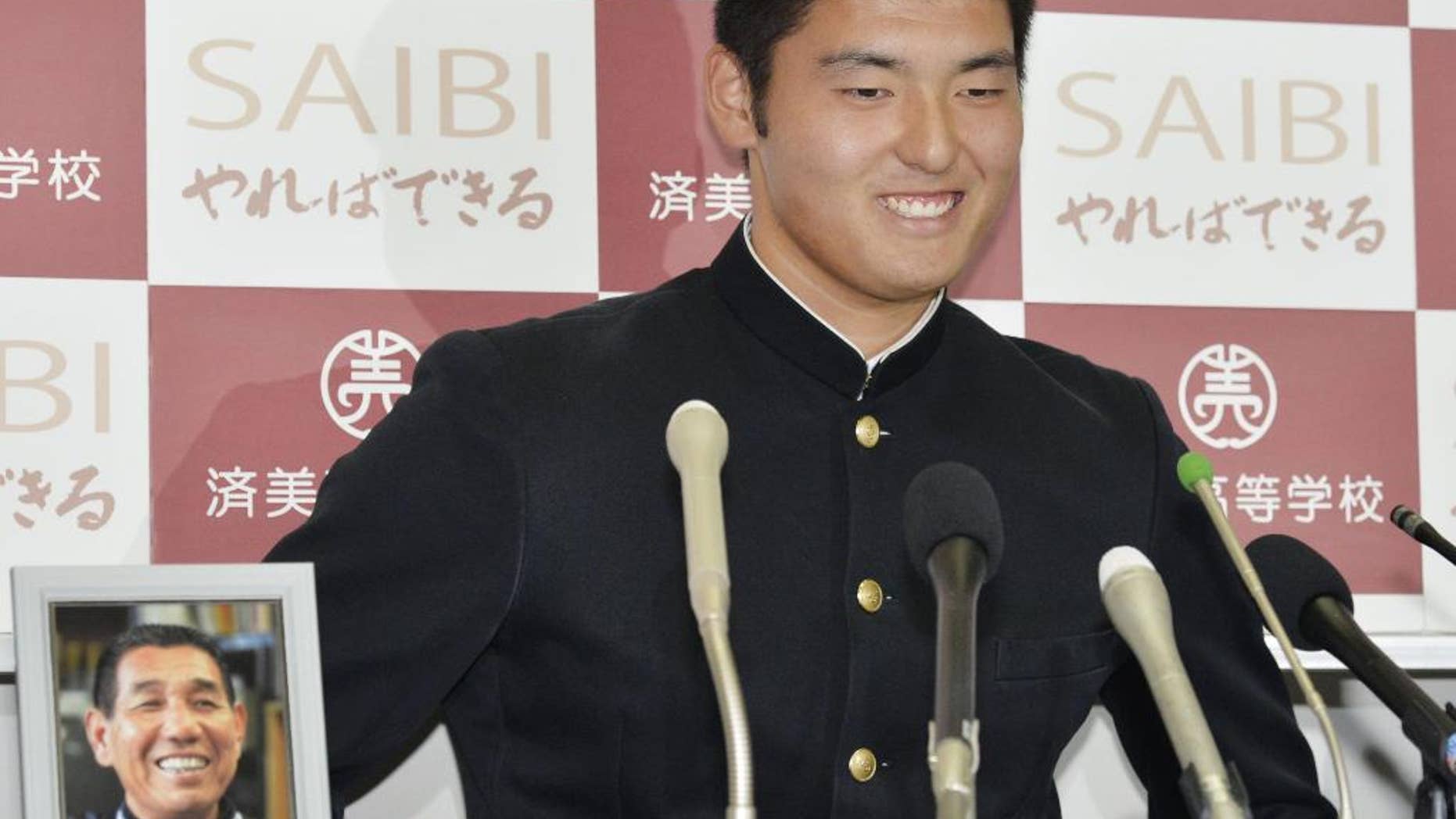 Japan's high school pitching star Tomohiro Anraku smiles next to a photo of his baseball team head coach Masanori Joko who died in September during a news conference at Saibi High School in Matsuyama, western Japan, Thursday, Oct. 23, 2014, after he was chosen first overall in Nippon Professional Baseball's amateur draft and was headed to the former team of New York Yankees pitcher Masahiro Tanaka. The Yakult Swallows and Rakuten Eagles selected Anraku, whose fastball has been clocked as high as 97 mph (156 kph), with the first and second picks respectively, and the Eagles won a lottery for the highly touted 17-year-old right-hander. (AP Photo/Kyodo News) JAPAN OUT, CREDIT MANDATORY