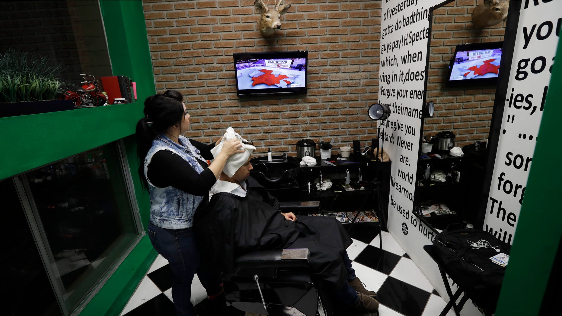 Paula Noriega, left, looks up at a screen displaying news of the elections in the United States as she dries the hair of Alan Maldonado at a barber shop Tuesday, Nov. 8, 2016, in Tijuana, Mexico. (AP Photo/Gregory Bull)
