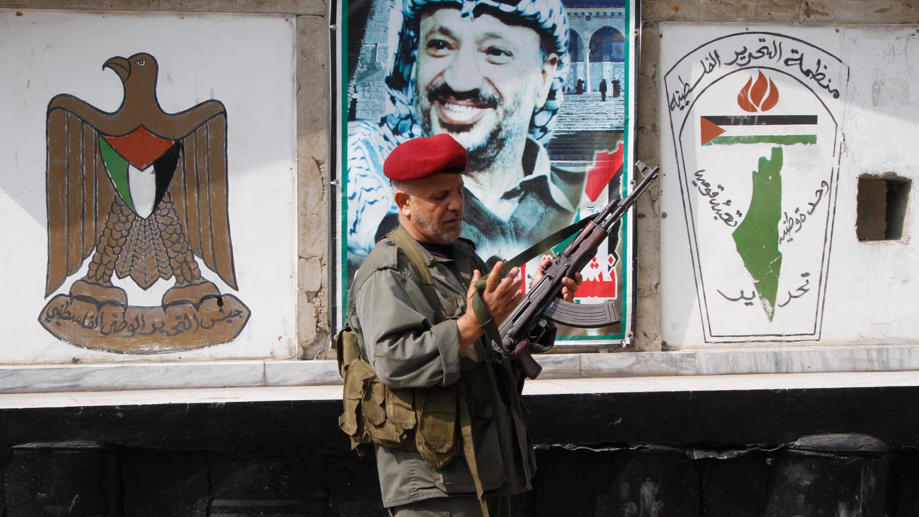 A member of the Palestine Liberation Organization's Armed Struggle unit, that is in charge of security in the Palestinian camps, holds his AK-47 machine gun as he walks in front of a portrait of the late Palestinian leader, Yasser Arafat, at a checkpoint in the Palestinian refugee camp of Ein el-Hilweh, in Sidon, Lebanon, Tuesday June 19, 2012. Palestinians refugees are protesting after clashes broke out Monday in the Nahr el-Bared camp in the north of Lebanon during the funeral of a Palestinian who was killed in a confrontation Friday. The Arabic logo painted on the wall, right, reads "The Palestine Liberation Organization, national unity, liberation." (AP Photo/Mohammed Zaatari)
