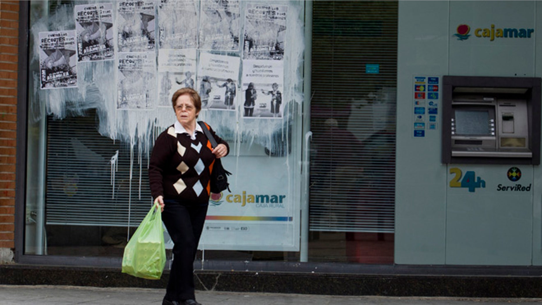 A woman leaves a bank plastered with protest posters in Madrid Monday May 21, 2012. (AP Photo/Paul White)