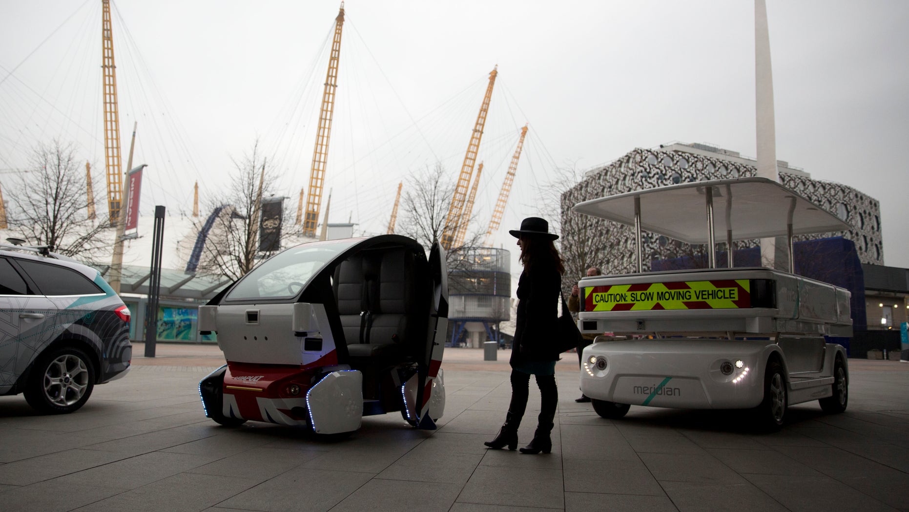 A woman poses for photographers beside a prototype driverless car called a LUTZ Pathfinder Pod, center, and a Meridian shuttle, right, during a launch event for the media near the O2 Arena in London.