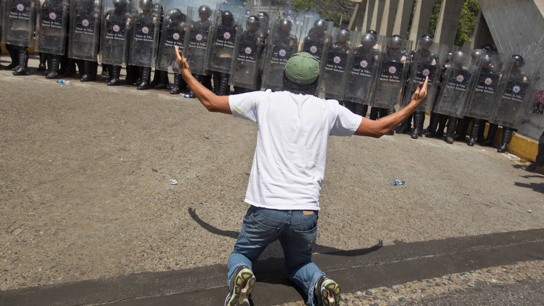 An anti-government demonsrator kneels in front of a formation of Bolivarian National Police in riot gear, during clashes at the Central University of Venezuela, UCV, in Caracas, Venezuela, Thursday, March 20, 2014. Thursday dawned with two more opposition politicians, San Cristobal Mayor Daniel Ceballos and San Diego Mayor Enzo Scarano, behind bars. Police used tear gas and water cannons to disperse a student-called protest of several thousand people in Caracas, some of those demonstrating against the arrests of the mayors. (AP Photo/Esteban Felix)