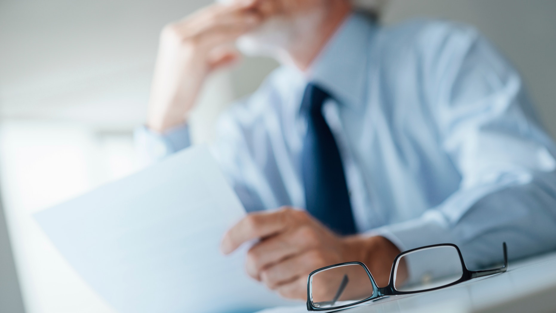 Pensive businessman with hand on chin looking away and holding a document, selective focus, glasses on foreground