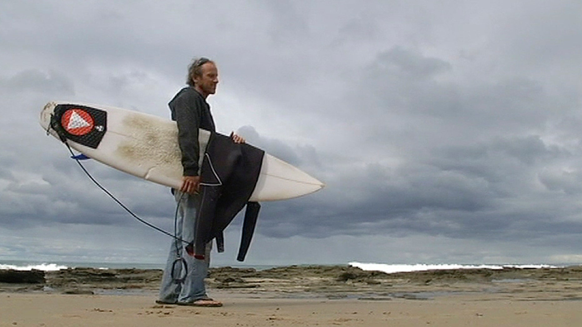 In this photo taken from video, Marcel Brunder, 37, holds his board as he looks out to sea after being attacked while surfing, Tuesday, Aug. 29, 2017, in the waters near Lorne, 140 kms. (90 miles) southwest of Melbourne, Australia. Surviving the attack with a scratch, torn wetsuit and teeth marks on his board, Brundler estimated the shark was a 3-meter (10-foot) great white shark. (Australia Broadcasting Corporation via AP)
