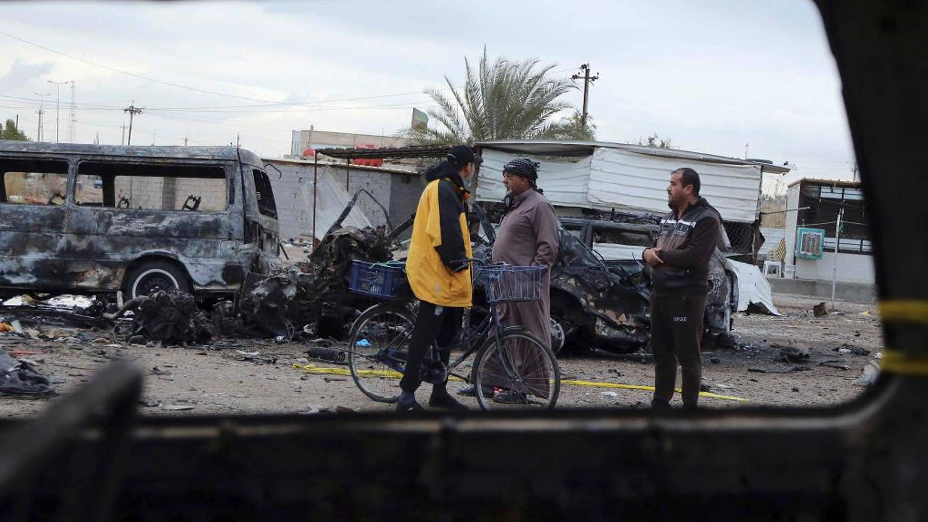 Civilians are seen through a broken window of a burned car at a used car dealer's parking lot in the southwestern al-Bayaa neighborhood, Baghdad, Iraq, Friday, Feb. 17, 2017, a day after a car bomb. The car bomb claimed by the Islamic State group killed scores of people. (AP Photo/ Karim Karim)