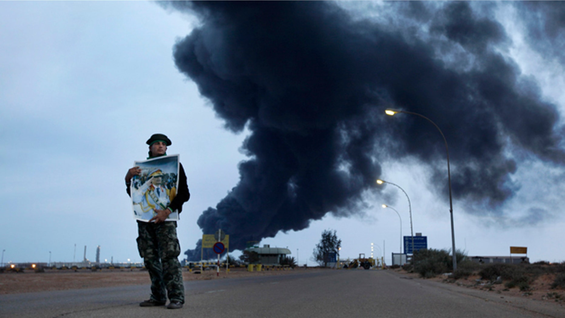 March 12, 2011: A pro-Gadhafi fighter holds a poster of Libyan Leader Moammar Gadhafi as black plume of smoke rises from the burning oil refinery as they are pictured during a government-organized visit for foreign media in Ras Lanouf, Libya