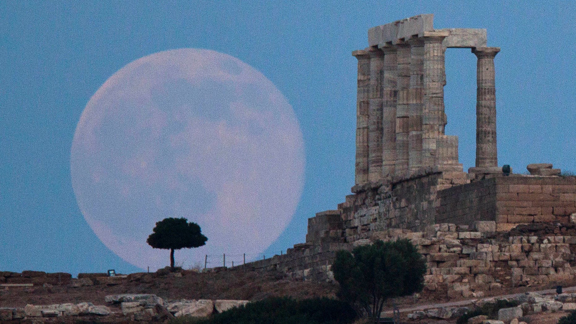 The full moon rises behind a tree next to the ruins of the ancient Temple of Poseidon near Athens, Greece.
