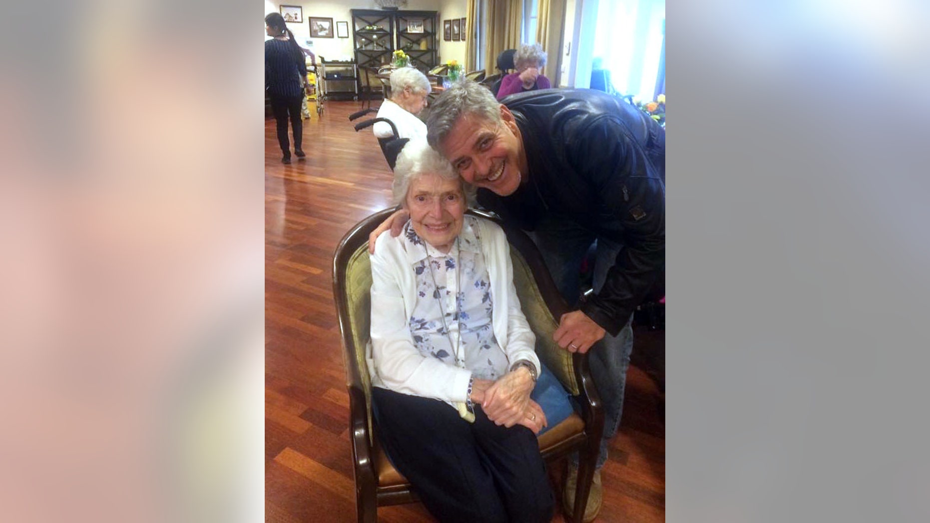 In this photo taken on Sunday, March 19, 2017, US actor George Clooney visits 87-year-old Pat Adams at Sunrise of Sonning Retirement and Assisted Living Facility in Reading, England. Actor George Clooney startled an 87-year-old fan in Britain by showing up at her assisted living facility with flowers and a card to wish her a happy birthday. (Linda Jones/Sunrise of Sonning Retirement and Assisted Living Facility via AP)