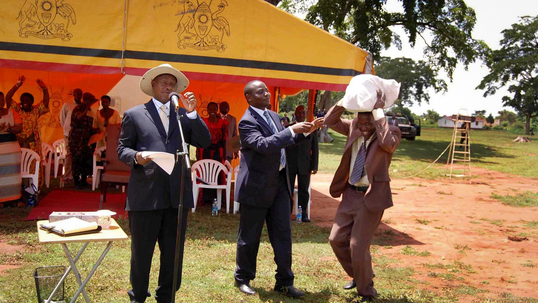In this photo taken on Saturday, April 20 2013 and released by the Uganda Presidency, Uganda's President Yoweri Museveni, left, and State Minister for Youth and Children Affairs Ronald Kibuule, center, hand over what the president's office said was a sack of money containing 250 million Ugandan shillings - about $100,000 - to an unidentified member of a partisan group of youths, right, in Kaliro district east of the capital Kampala, in Uganda. It’s become an infamous photo in Uganda. The country’s leader looks on in bemusement as a young man bends low to carry a sack filled with a donation from the president _ almost $100,000 in cash. The money is a donation from President Yoweri Museveni to a partisan group of youths in eastern Uganda, where he struggles to win votes in national elections, and activists and opposition politicians are seizing upon the moment as a blatant example of political corruption in Uganda. The picture, which was distributed by the president’s office after the event last weekend, was meant to highlight Museveni’s generosity toward a group of youth. Instead it has focused attention on Museveni’s profligacy during political tours and what some say is his role in fueling graft in the East African country that he has led since 1986. (AP Photo/Uganda Presidency)