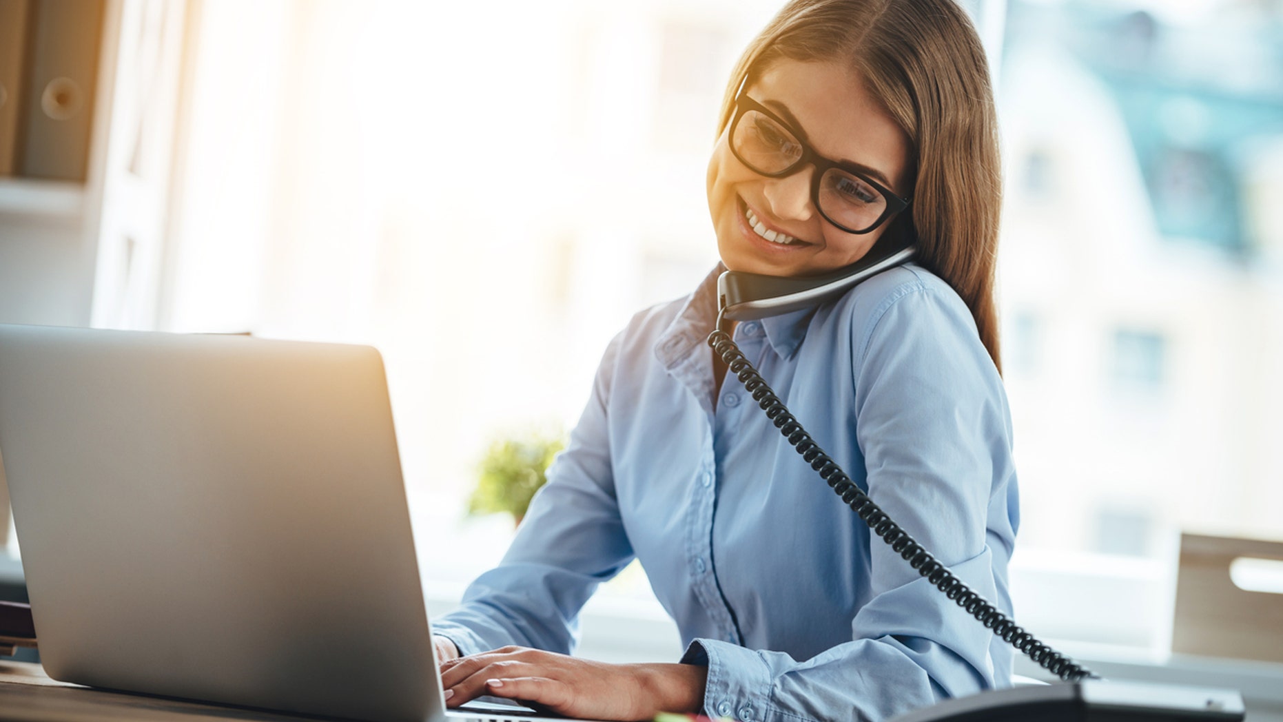 Cheerful young beautiful woman in glasses talking on the phone and using laptop with smile while sitting at her working place