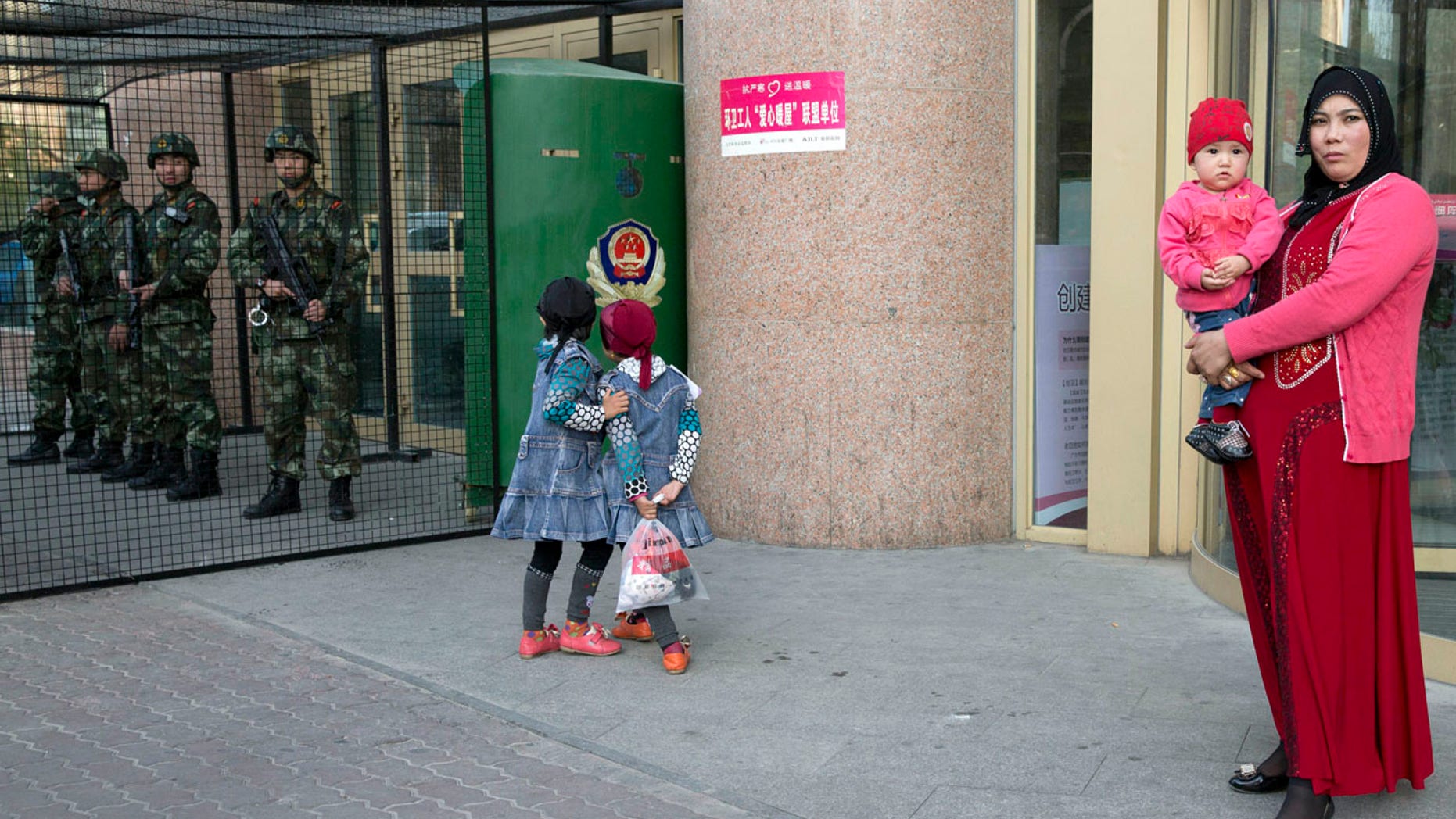 In this Thursday, May 1, 2014 file photo, an Uighur woman carries a toddler as children play near a cage protecting heavily armed Chinese paramilitary policemen on duty in Urumqi in northwest China's Xinjiang Uygur Autonomous Region.