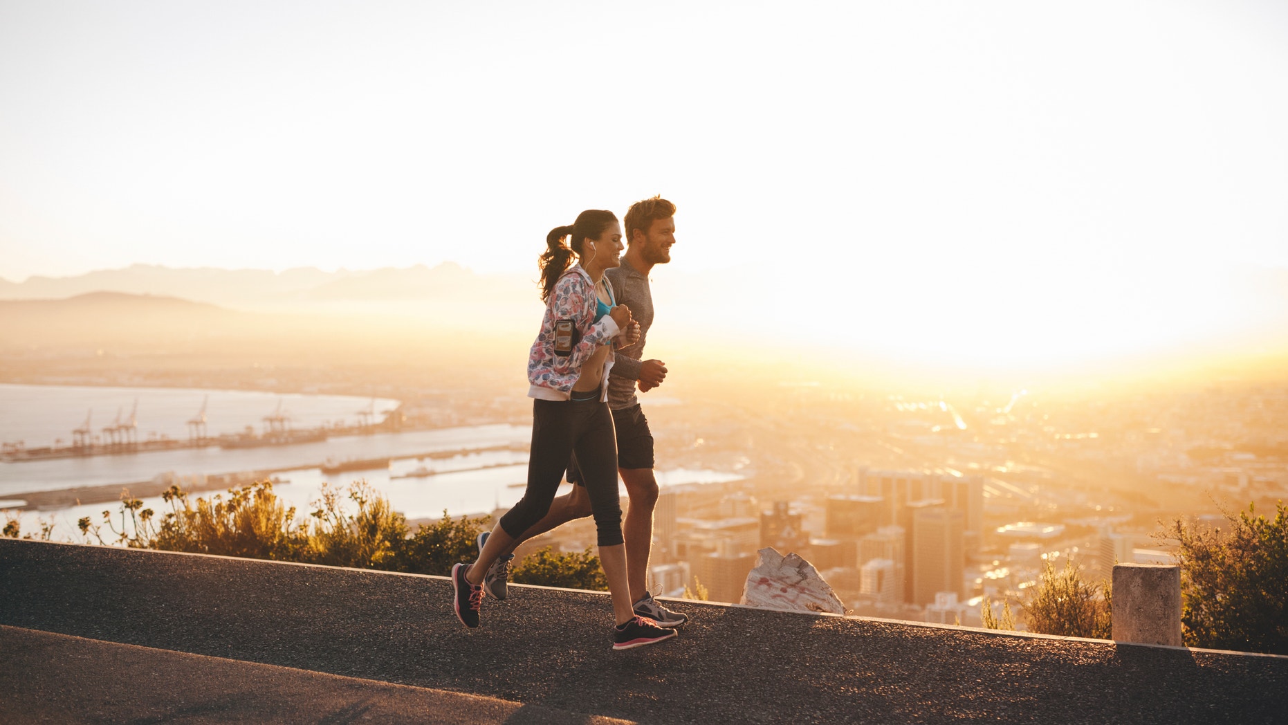 Shot of young couple running on hillside road outside the city. Young man and woman jogging in morning with bright sunlight.