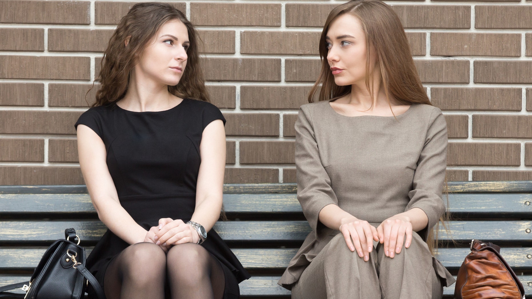 Portrait of two beautiful young female rivals sitting side by side on bench and looking at each other with challenging expressions. Attractive caucasian office women ready for confrontation