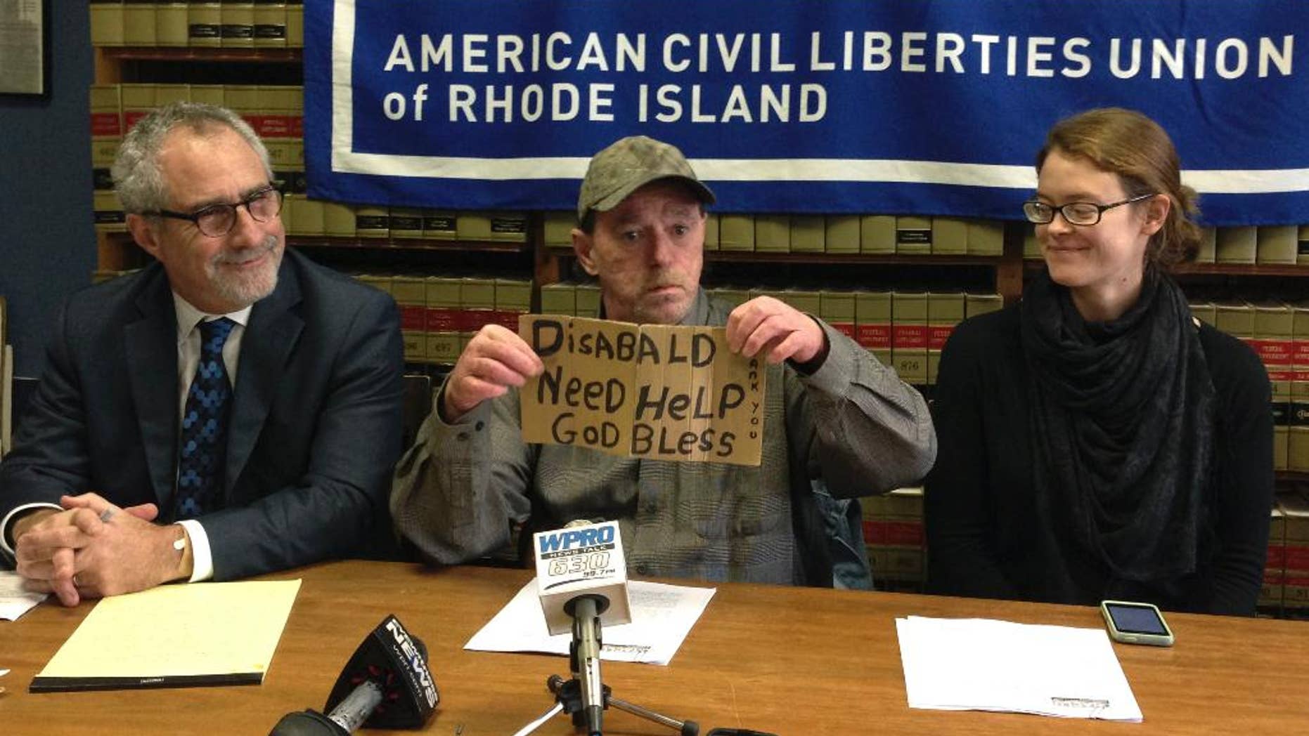 Michael Monteiro, center, with his lawyer Marc Gursky, left, and outreach worker Megan Smith, right, holds the sign that he used to solicit donations from a median strip in Cranston, R.I., during a news conference Wednesday, Dec. 16, 2015 in Providence, R.I. Monteiro said he has periodically asked for donations at the intersection for years, but stopped this summer after he was cited by police, then told by a judge not to return. Monteiro is now suing the city for violating his constitutional rights. (AP Photo/Michelle R. Smith)