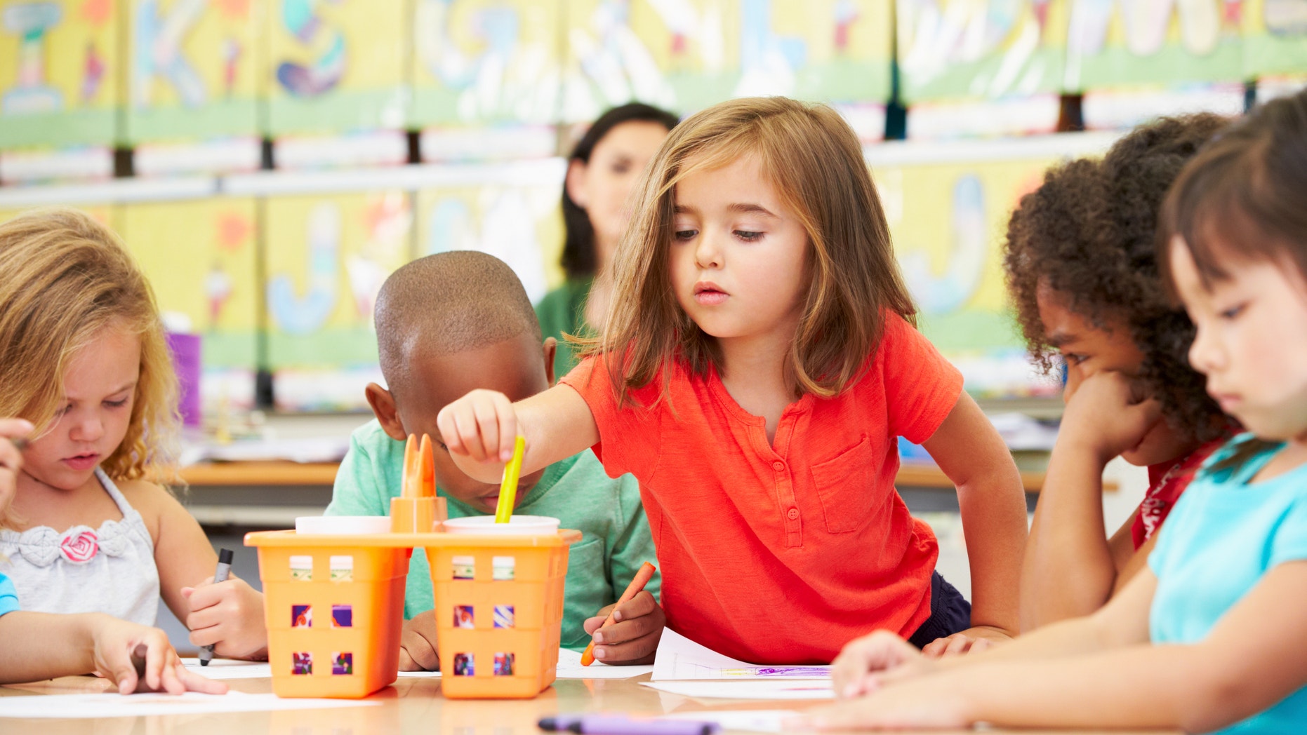 Group Of Elementary Age Children In Art Class With Teacher Reaching For Crayon