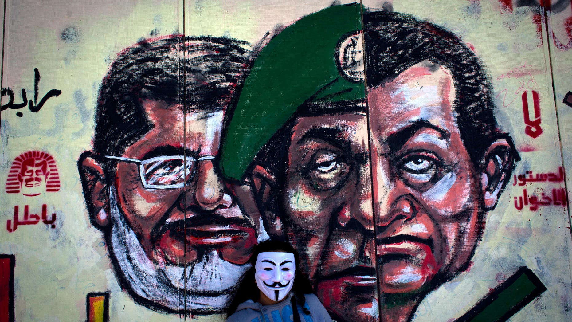 FILE - In this Dec. 8, 2012 file photo, an Egyptian protester wears a Guy Fawkes mask while posing for a photo next to a mural painted overnight on the exterior wall of the presidential palace depicting president Mohammed Morsi, left, former military council ruler Hussein Tantawi, center and ousted President Mubarak with Arabic that reads "No, the brotherhood's constitution is not valid, in Cairo. Egyptian authorities switched the venue for the trial of the former Islamist president on Sunday, Nov. 3, 2013 a last-minute change made after the Muslim Brotherhood called for mass demonstrations at the original location. The trial of Morsi, now to be held east of the capital on Monday, could lead to another round of bloodshed as his supporters look likely to face an emboldened security apparatus that has boosted its forces for the hearing.(AP Photo/Nasser Nasser, File)