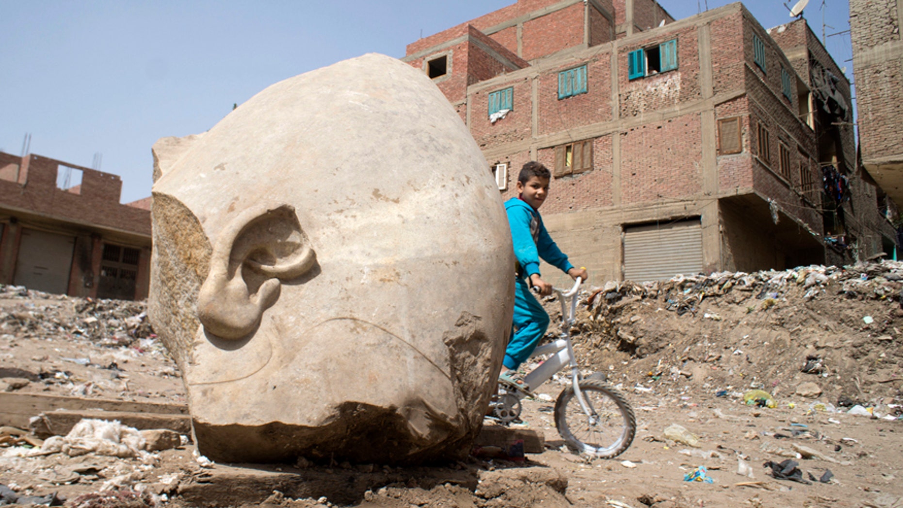 A boy rides his his bicycle past a recently discovered statue in a Cairo slum that may be of pharaoh Ramses II, in Cairo, Egypt, Friday, March 10, 2017. Archeologists in Egypt have discovered a massive statue that may be of pharaoh Ramses II, one of the country's most famous ancient rulers. The colossus, whose head was pulled from mud and groundwater by a bulldozer on Thursday, is around eight meters (yards) tall and was discovered by a German-Egyptian team. (AP Photo/Amr Nabil)