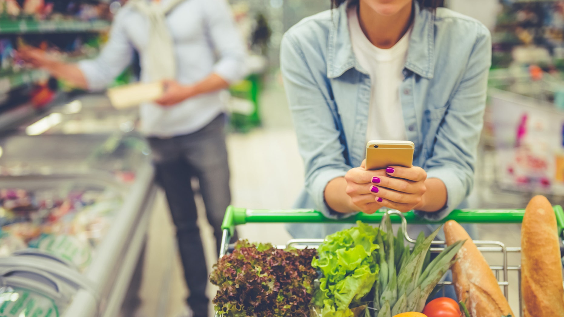 Couple in the supermarket. Cropped image of girl leaning on shopping cart, using a mobile phone and smiling, in the background her boyfriend is choosing food