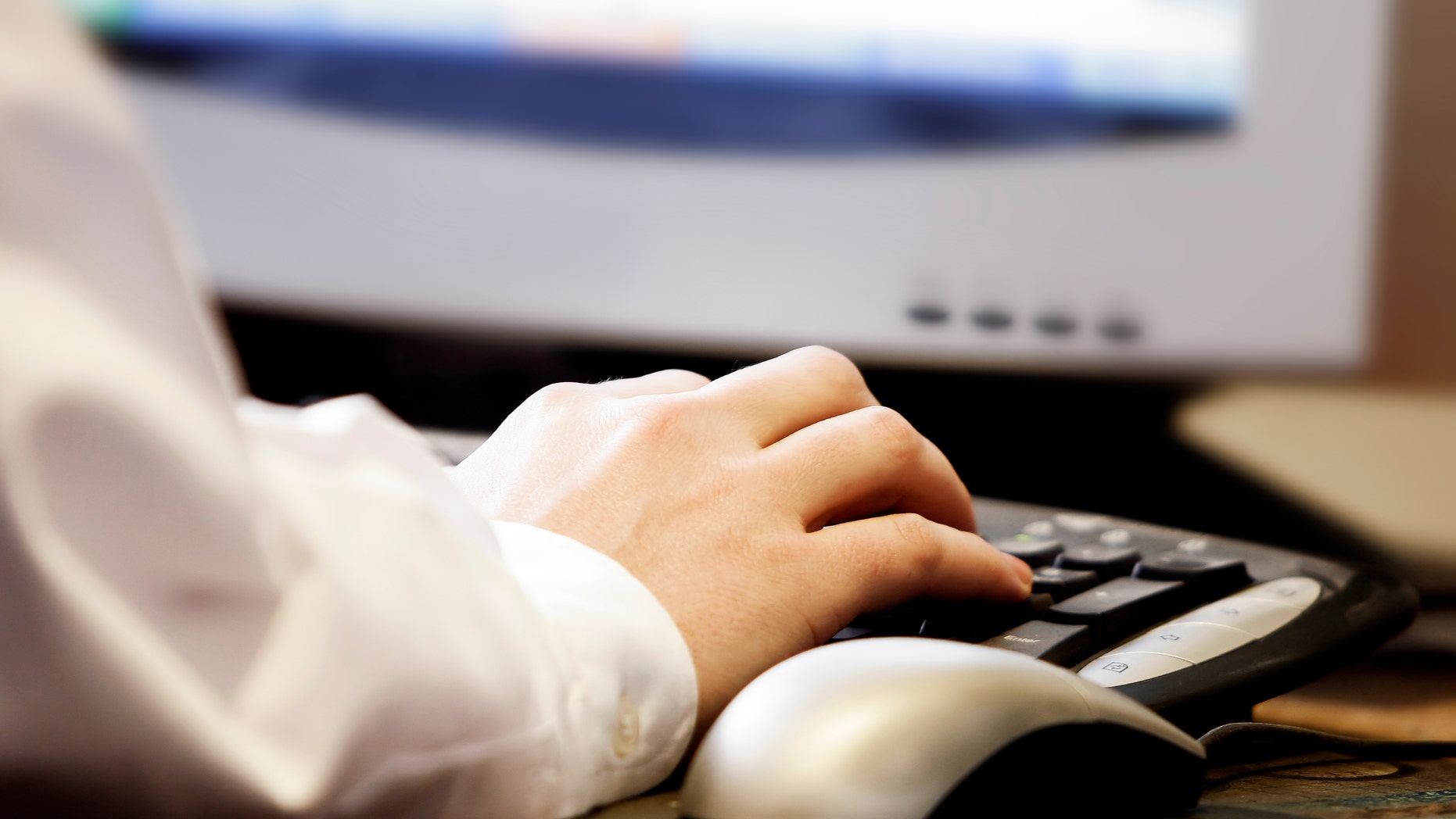 Close-up of a man's hand typing on a computer keyboard, with monitor and mouse (shallow focus).