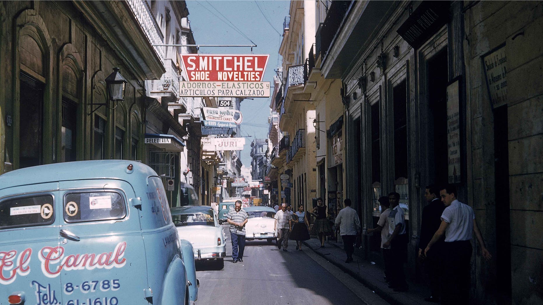 Cars are parked along a narrow street as pedestrians walk in the shade in Havana, Cuba, 1950s.