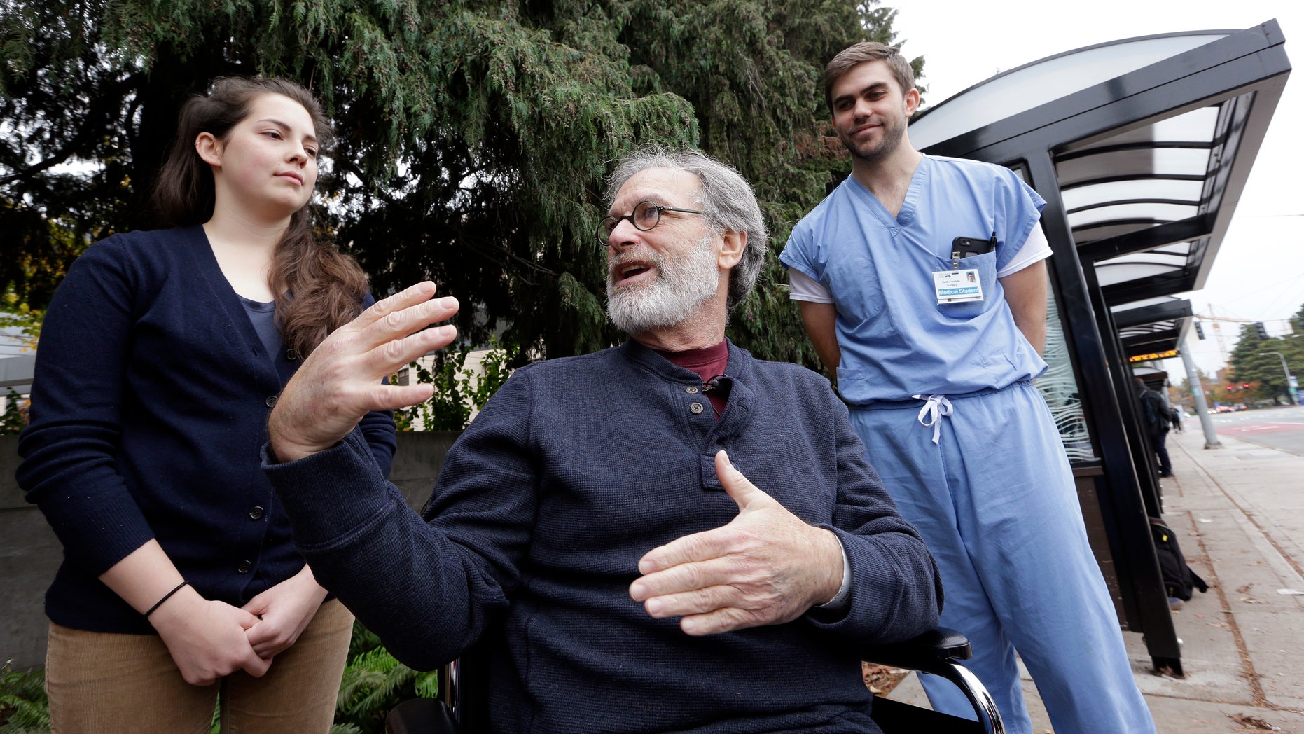 Stephen DeMont, center, sits in a wheelchair at the bus stop where he says that Zach Forcade, right, and Madeline Dahl saved his life days earlier in front of the University of Washington Medical Center, Wednesday, Oct. 19, 2016, in Seattle. When DeMont collapsed at a bus stop in front of the UW Medical Center on his morning commute, Dahl was one of 41 people within a 330-yard radius who happened to have a cell phone app alerting them to the emergency. Forcade, a medical student, witnessed the collapse and rushed over to begin chest compressions, as within moments Dahl, a cardiac nurse just getting off her shift in the hospital, was alerted by her phone and sprinted down the sidewalk, assisting until paramedics arrived. (AP Photo/Elaine Thompson)