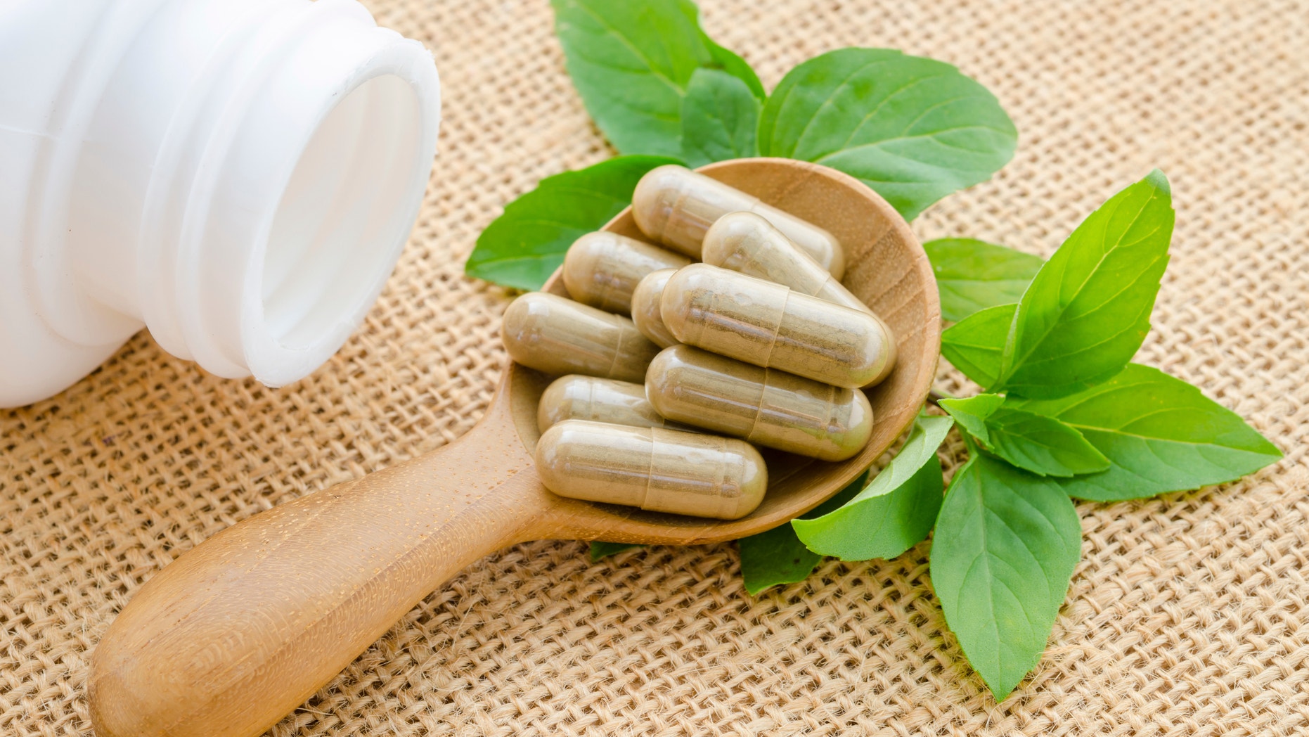 Herb capsule spilling out of a white bottle in wooden spoon with green leaf on sack background.